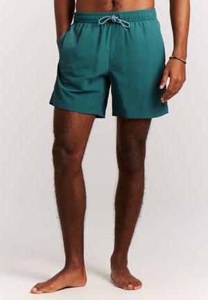 Modèle masculin portant un short de bain turquoise avec taille à cordon, debout pieds nus, les mains détendues le long du corps, sur un fond uni.
