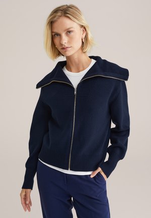 Maglione zip-up blu navy con colletto a coste ampie, tessuto a maglia testurizzato e zip argentata. Indossato sopra una camicia bianca, abbinato a pantaloni blu navy.