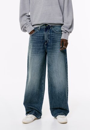 Persoon in loszittende vervaagde blauwe jeans, lichtgrijze sweatshirt, witte sneakers, met één hand in de zak en een zilveren ring om de andere hand.