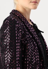 Veste noire ornée de sequins de différentes tailles et nuances de rose, dotée d'un col et d'une fermeture à cravate à l'avant. Le tissu texturé met en valeur le design.