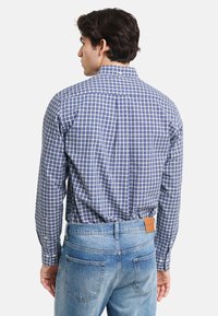 Camisa de manga larga a cuadros en azul y blanco, diseño ligeramente ajustado, material de algodón, puños abotonados y detalle de yugo en la parte posterior, combinada con vaqueros azul desgastados.