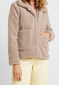 Fleecejakke i lys beige med krave-design, to forlommer og lange ærmer. Blød tekstur med synlige stikninger.