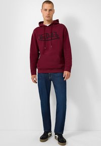 Felpa cappuccio marrone con logo nero "Von Dutch", cordini, tasca frontale; abbinata a jeans blu scuri e sneakers nere. Texture del tessuto liscia.