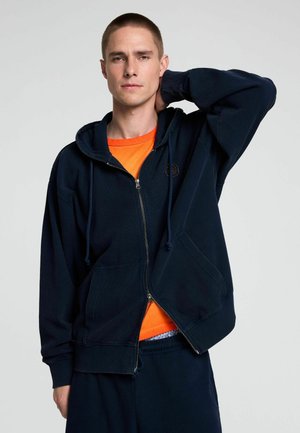 Mladý muž s krátkými vlasy, který má na sobě tmavě modrou mikinu na zip přes oranžové tričko, pózující s jednou rukou za krkem.