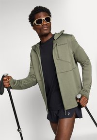 Giacca zip-up verde oliva con un motivo testurizzato, t-shirt nera e pantaloni corti scuri; indossa occhiali da sole beige e tiene un bastone da trekking.