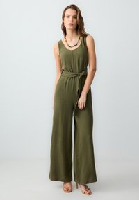 Grenouillère vert olive avec un décolleté arrondi, des jambes larges et une ceinture à nouer à la taille. Comprend deux poches avant et un tissu texturé.
