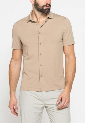 Uomo che indossa una camicia beige a maniche corte abbottonata e pantaloni chiari, guarda verso sinistra su uno sfondo neutro.