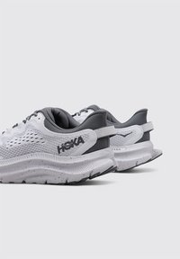 Scarpe da ginnastica grigie e bianche con tomaia in mesh traspirante, colletto imbottito e suola spessa e texturizzata con macchie. Branding HOKA sul lato.