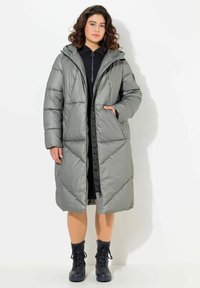 Donna che indossa un lungo cappotto imbottito grigio con cappuccio, stivali neri stringati e un abito nero, in piedi contro uno sfondo bianco.