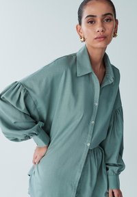 Light green oversized button-up shirt with billowy sleeves, a soft texture, and a relaxed fit, featuring a collared neckline and front pockets.