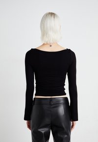 Svart langermet topp med en tettsittende design, rynket tekstur på baksiden og rund halskant. Kombinert med høyhåndte svarte skinnbukser.