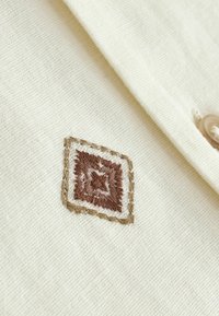 Cremfarbener Stoff mit einem aufgenähten quadratischen Stück, das ein braun-beiges geometrisches Muster in der Nähe eines geknöpften Kragenrandes zeigt.