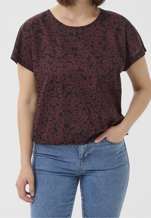 Femme portant un haut à manches courtes rouge foncé à motif floral et un jean bleu, une main dans la poche, sur un fond uni.