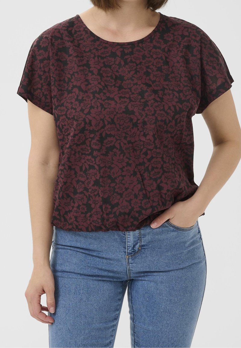 Femme portant un haut à manches courtes rouge foncé à motif floral et un jean bleu, une main dans la poche, sur un fond uni.