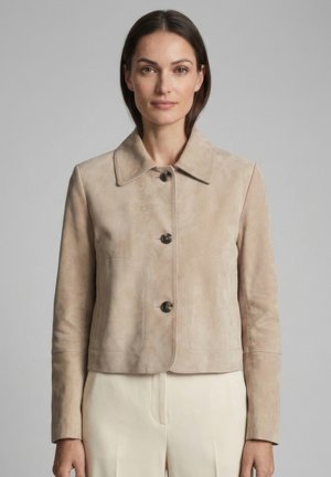 Frau trägt eine beiger Wildleder-Kurjacke mit drei großen Knöpfen, langen Ärmeln und Kragen, stehend vor einem schlichten Hintergrund.