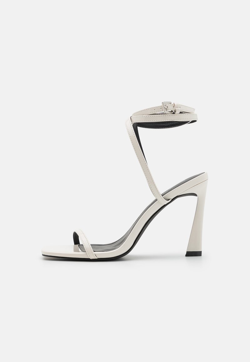 Λευκές παλαιές δερμάτινες ψηλοτάκουνες σανδάλες με κλειστή φτέρνα, αυτοκόλλητο που φτάνει μέχρι τον αστράγαλο και τετράγωνη μύτη. Διαθέτουν λεία υφή και λεπτό τακούνι.