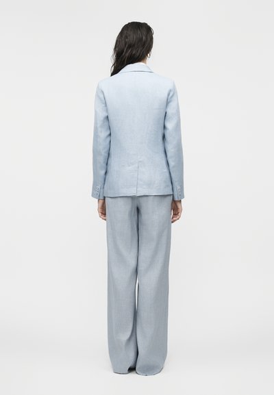 Γυναίκα με μακριά σκούρα μαλλιά που φοράει ένα ανοιχτό γαλάζιο σακάκι με κοψίματα και παντελόνι με φαρδιά μπατζάκια, στέκεται με την πλάτη στραμμένη σε απλό φόντο.