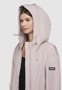 Svetloružová bunda s kapucňou z ľahkého materiálu. Obsahuje šnúrky na stiahnutie, zips a nášivku s logom na ľavom rukáve.