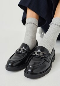 Black leather loafers with a chunky silver chain accent, raised stitching, and a thick rubber sole. Worn with light gray ribbed socks.