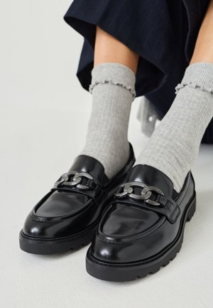 Zwarte leren loafers met een chunky zilveren kettingaccent, verhoogde stiksels en een dikke rubberen zool. Gedeeltelijk gedragen met lichtgrijze geribbelde sokken.