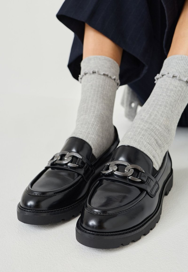 Black leather loafers with a chunky silver chain accent, raised stitching, and a thick rubber sole. Worn with light gray ribbed socks.