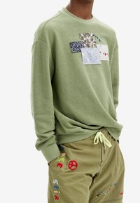 Felpa verde oliva con design a patchwork, caratterizzata da quattro quadrati di tessuto multicolore, finitura testurizzata e polsini a coste.