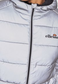 Doudoune gris clair avec fermeture éclair noire et logo ellesse sur la poitrine gauche, portée par une personne avec cou et menton visibles.