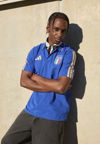 Μπλε polo μπλούζα με χρυσές λεπτομέρειες και έμβλημα της Ιταλίας, συνδυασμένη με σκούρο γκρι τζιν. Λείο ύφασμα και άνετη εφαρμογή.