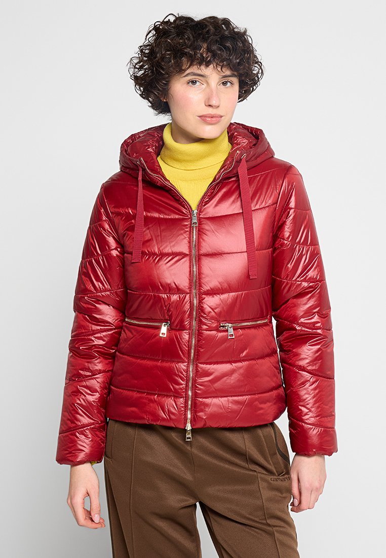 Persoon met kort, krullend haar die een glanzende rode pufferjack over een gele turtleneck en een paarse broek draagt, staande tegen een effen achtergrond.