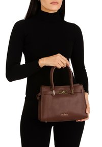 Vrouw in een zwart langemouwen top die een gestructureerde donkerbruine leren handtas vasthoudt met gouden accenten en bovenhandvatten.