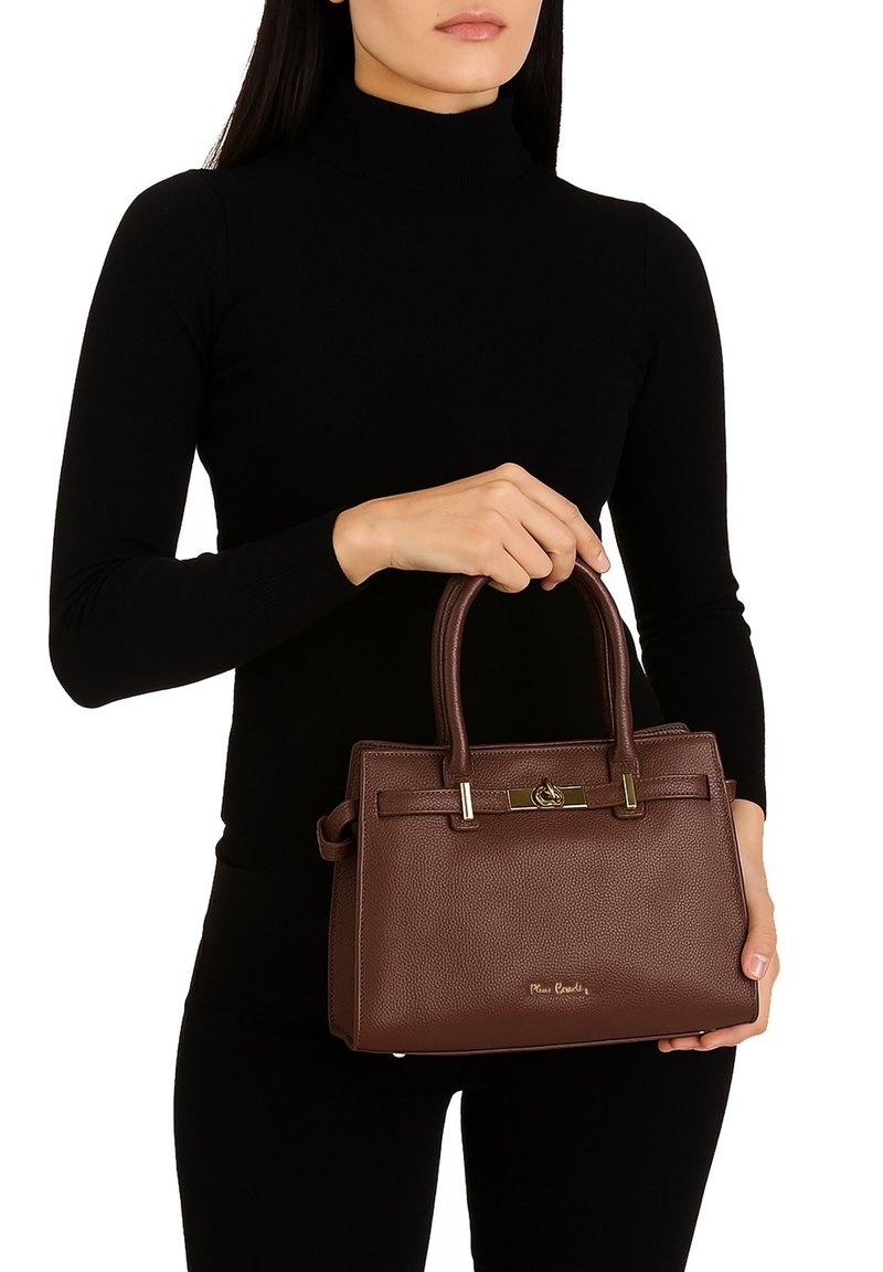 Vrouw in een zwart langemouwen top die een gestructureerde donkerbruine leren handtas vasthoudt met gouden accenten en bovenhandvatten.