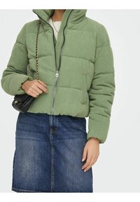 Chaqueta acolchada de pana verde con capucha, con cremallera frontal, textura acanalada y dobladillo elástico, combinada con una falda vaquera.