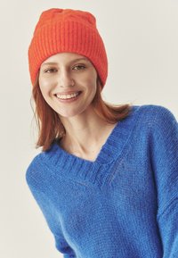 Niebieski, teksturowany sweter z dekoltem w serek, zestawiony z jasnopomarańczową, prążkowaną czapką. Materiały wydają się miękkie i ciepłe.