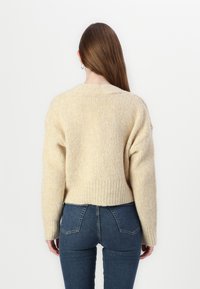 Pull en tricot de couleur crème, avec une coupe décontractée, présentant une texture côtelée et une longueur cropped, associé à un jean skinny bleu foncé.