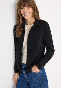 Giacca nera con zip e colletto alto, con dettagli testurizzati sulle maniche, indossata sopra una maglietta beige chiaro e jeans blu.