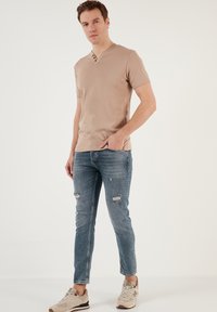 Béžové tričko s krátkým rukávem a výstřihem do V se třemi knoflíky, kombinované se světle modrými slim-fit džínami se vybledlými detaily a světlými teniskami.