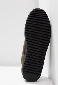 Sohle eines dunkelbraunen Schuhs mit dicken, tiefen horizontalen Rillen, aufrecht an einer weißen Wand- und Bodenecke lehnend.