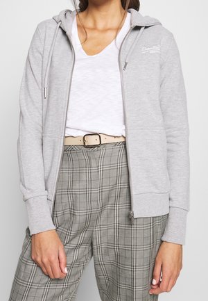 Vrouw draagt een lichtgrijze hoodie met ritssluiting, een wit V-hals T-shirt, een beige riem en grijze geruite broek, staand met ontspannen armen.