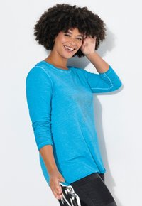 Türkisfarbenes Langarmshirt mit weicher Textur und einem großen Strassstern-Design auf der Vorderseite, kombiniert mit dunklen Skinny-Jeans.