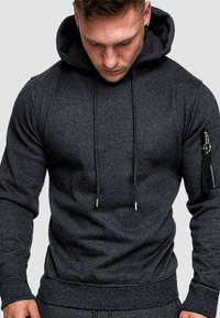 Dunkelgrauer Kapuzenpullover aus strukturiertem Stoff, mit Kordelzügen und einer Reißverschlusstasche am linken Ärmel. Figurbetonter Schnitt.