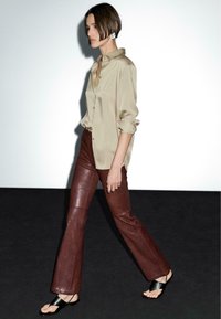 Camicia di satin beige, pantaloni in pelle marroni a zampa d'elefante e sandali neri scivolati. Modella cammina su uno sfondo neutro e con illuminazione minimale.