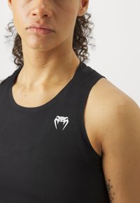 Čierna športová tielko s textúrovanou látkou. Má biele logo stylizovanej pavúka blízko výstrihu.