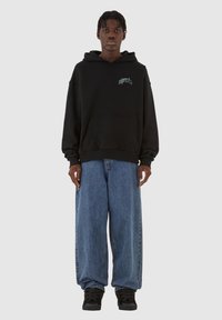 Svart hettegenser med frontlomme, teksturert stoff og brodert design. Kombinert med løstsittende blå denimjeans og svarte joggesko.