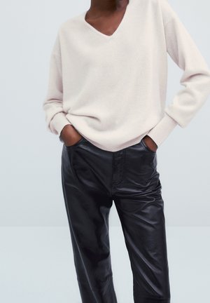 Personne portant un pull col V crème et un pantalon en cuir noir, les mains dans les poches, sur un fond clair uni.