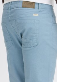 Lichtblauwe denim broek met een gestructureerde afwerking, twee achterzakken met schuine kleppen en een bruine label op de tailleband.