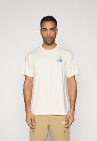 Μπέζ βαμβακερό μπλουζάκι με κοντά μανίκια και μπλε λογότυπο στο αριστερό στήθος. Απλό σχέδιο με στρογγυλή λαιμόκοψη.