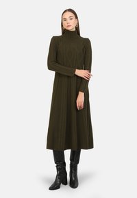 Gebreide jurk in olijfgroen met een hoge hals, voorzien van een textuurpatroon en lange mouwen. Gekleed met zwarte kniehoge laarzen.