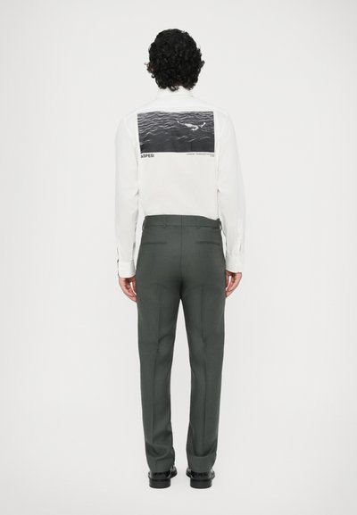 Balta krekla ar grafisku druku uz muguras, kopā ar piegulošām tumši zaļām biksēm un melniem apaviem; gluda auduma un strukturēta piegriezuma.