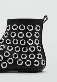 Botas de tobillo de cuero negro con grandes ojales plateados dispuestos en un patrón de rejilla. El diseño incluye una pestaña para ayudar a ponerlas en la parte posterior.