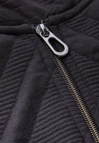 Haut à capuche noir zippé avec un motif texturé. Comprend une fermeture éclair métallique et un onglet de traction arrondi, mettant en avant un design moderne et épuré.